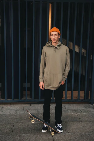 Skateboarder en sudadera con capucha de gran tamaño con tabla de pie delante de la valla de metal - foto de stock