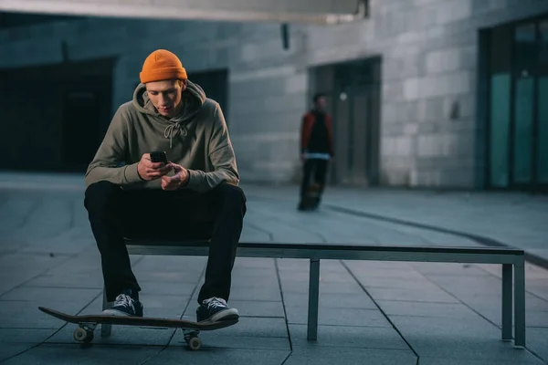 Skateboarder en ropa de calle moderna sentado en el banco y utilizando el teléfono inteligente - foto de stock