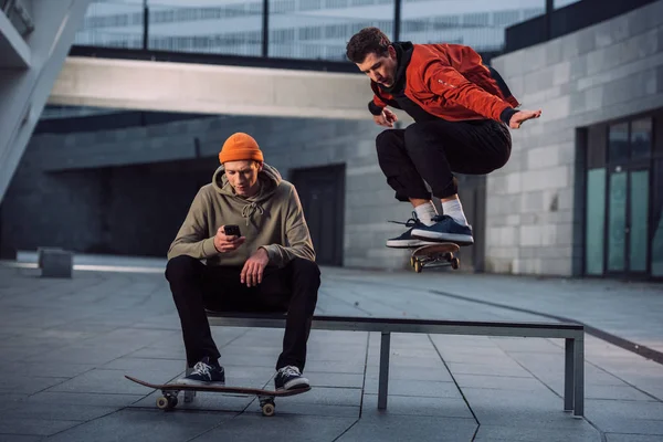 Skateboarder jumping over bench while his partner sitting on it and using phone — Stock Photo