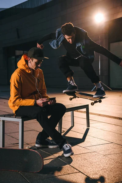 Skateboarder jumping over bench while man sitting on it and listening music — Stock Photo