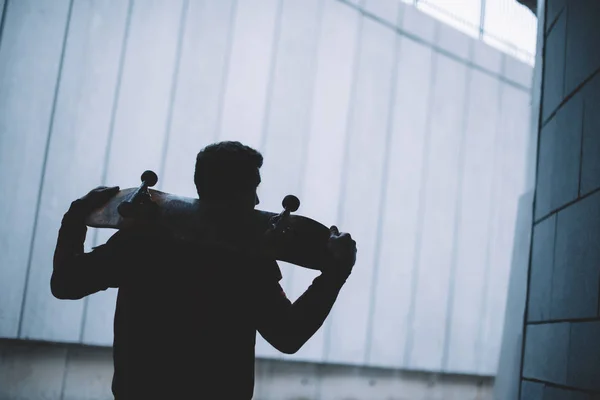 Skateboarder giovane in piedi con tavola sulle spalle davanti al muro di cemento grigio — Foto stock