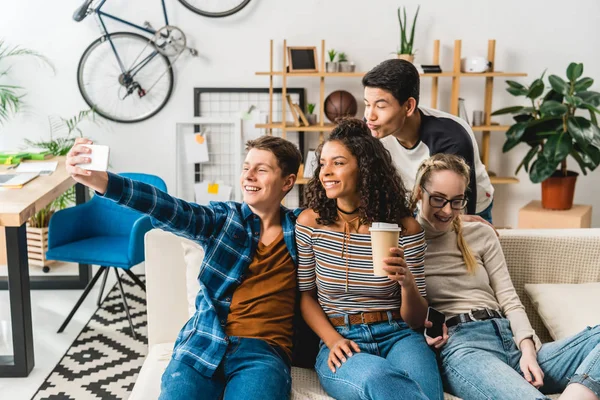 Happy multiethnic teenagers sitting on sofa and taking selfie with smartphone — Stock Photo