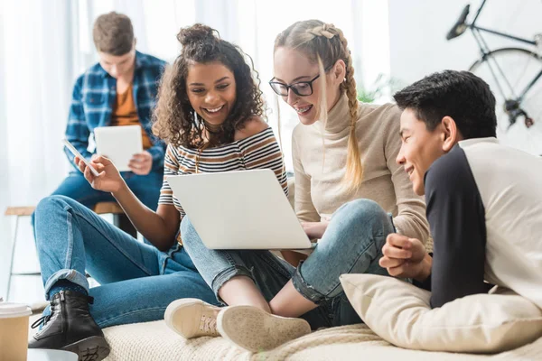 Adolescentes multiculturales felices mirando a la computadora portátil y sentado en el sofá - foto de stock