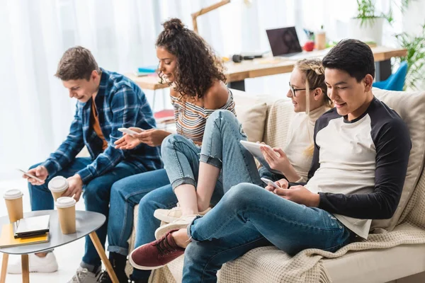 Happy multicultural teens sitting on sofa with digital devices — Stock Photo