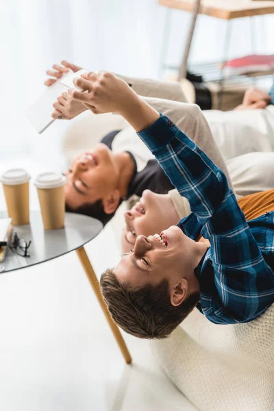 Caucasian teenagers lying on sofa and taking selfie with tablet and smartphone — Stock Photo