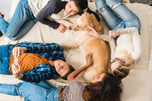 Visão aérea de adolescentes multiétnicos deitados na cama e tomando selfie com o cão — Fotografia de Stock