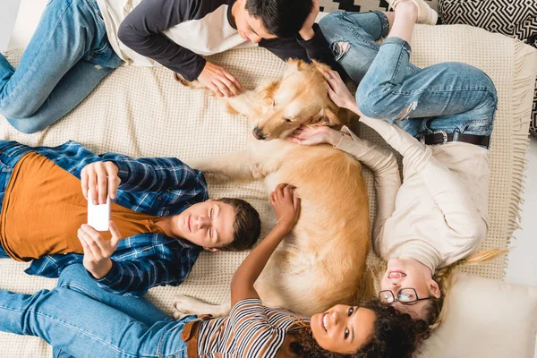 Vista aérea de adolescentes multiculturales acostados en la cama y tomando selfie con el perro - foto de stock