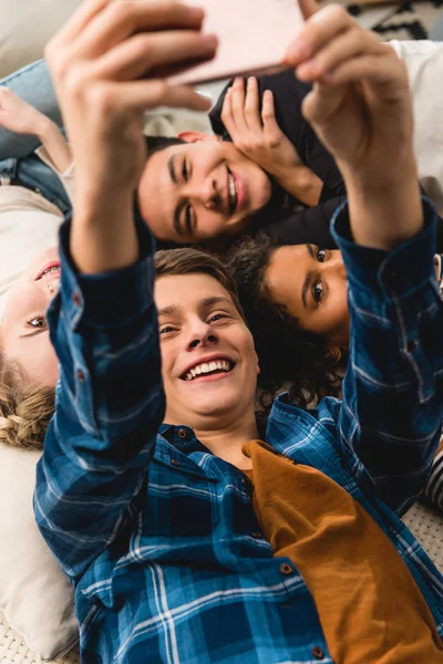 Vista aérea de adolescentes multiculturais tomando selfie enquanto deitado na cama — Fotografia de Stock