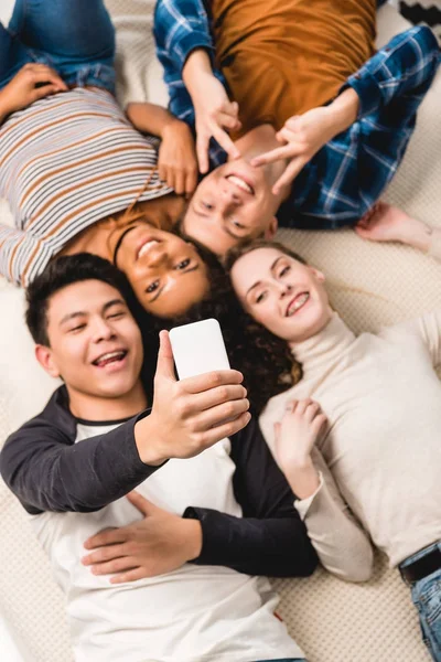 Vista aérea de adolescentes multiétnicos tomando selfie mientras están acostados en la cama - foto de stock