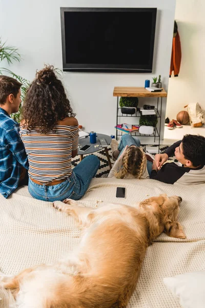 Four teens watching tv and dog lying on bed — Stock Photo