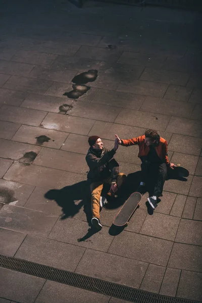 Vista de ángulo alto de los patinadores que dan cinco altos mientras están sentados en el suelo - foto de stock