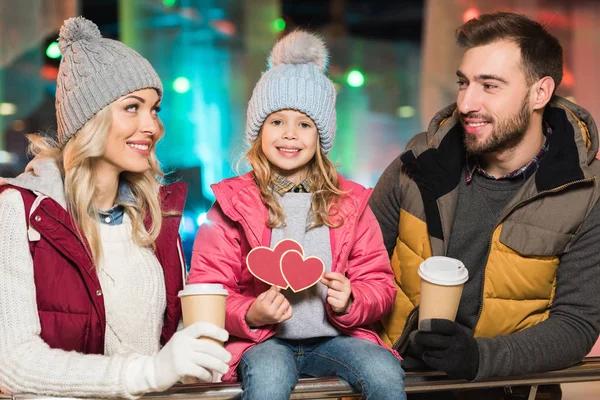Parents heureux avec café pour aller regarder mignonne petite fille tenant des coeurs symbole sur la patinoire — Photo de stock