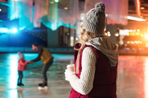 Young woman drinking coffee from paper cup and looking at happy family skating behind on rink — Stock Photo