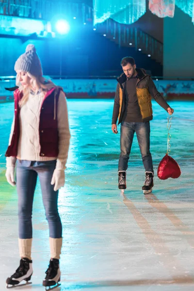Jeune femme debout sur la patinoire tandis que petit ami avec ballon en forme de coeur debout derrière — Photo de stock