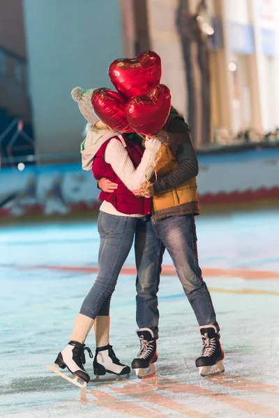 Heureux jeune couple avec des ballons en forme de coeur étreignant sur la patinoire à st Saint-Valentin — Photo de stock