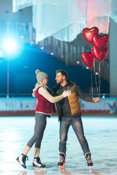 Schönes glückliches junges Paar mit herzförmigen Luftballons, die einander auf der Eisbahn lächeln — Stockfoto