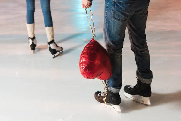 Plan recadré de jeune homme tenant ballon en forme de coeur tandis que la petite amie s'en va sur la patinoire — Photo de stock