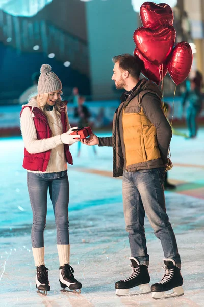 Vue latérale du jeune homme avec des ballons présentant boîte cadeau à petite amie à la Saint-Valentin st sur la patinoire — Photo de stock