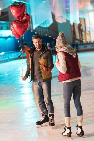 Fröhlicher junger Mann mit Luftballons überreicht Freundin am Valentinstag auf der Eisbahn Geschenkbox — Stockfoto