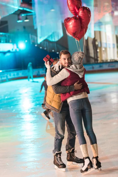 Vue pleine longueur de la jeune femme tenant boîte cadeau et étreignant petit ami heureux avec des ballons sur la patinoire — Photo de stock