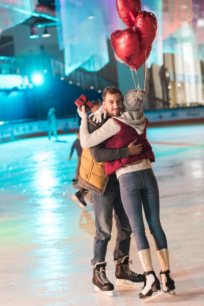 Vista completa de la mujer joven sosteniendo caja de regalo y abrazando novio feliz con globos en la pista - foto de stock