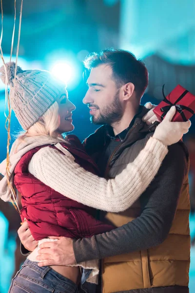 Hermosa feliz joven pareja con caja de regalo mirándose en pista de patinaje - foto de stock