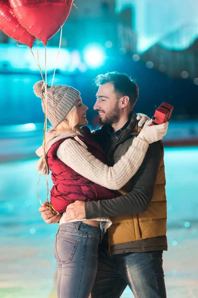 Heureux jeune couple avec boîte-cadeau et ballons en forme de coeur se regardant sur la patinoire — Photo de stock