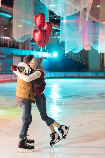 Seitenansicht eines glücklichen jungen Paares mit Geschenkbox und herzförmigen Luftballons, die sich auf der Eisbahn umarmen — Stockfoto
