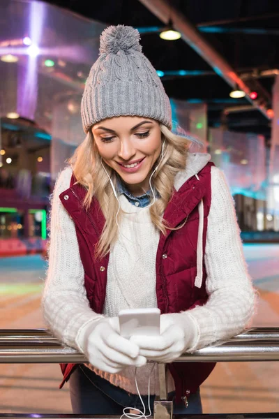 Belle jeune femme souriante dans les écouteurs en utilisant smartphone sur la patinoire — Photo de stock