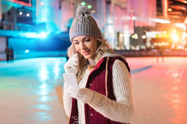 Lächelnde junge Frau hält Smartphone in der Hand und hört Musik in Kopfhörern auf der Eisbahn — Stockfoto