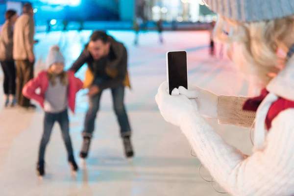 Cropped shot of woman holding smartphone with blank screen and photographing family skating on rink — Stock Photo