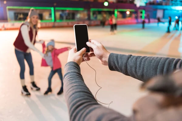 Cropped shot of man holding smartphone with blank screen and photographing family skating on rink — Stock Photo
