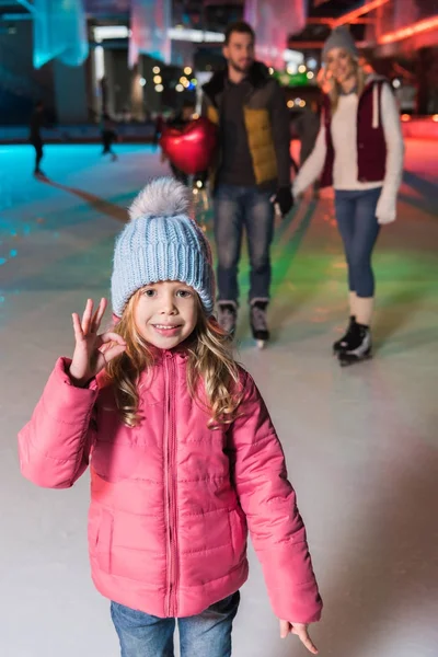 Bonito menina mostrando ok sinal e sorrindo para a câmera enquanto patinação no gelo com os pais no ringue — Fotografia de Stock