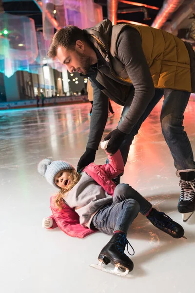 Padre aiutare piccola figlia sdraiata sul ghiaccio sulla pista di pattinaggio — Foto stock