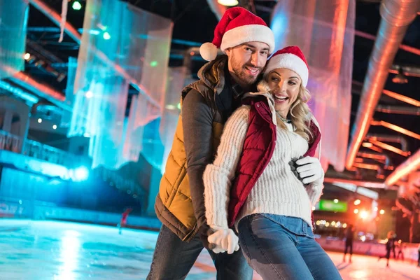 Bella felice giovane coppia in cappelli di Babbo Natale abbracciare e divertirsi sulla pista di pattinaggio — Foto stock