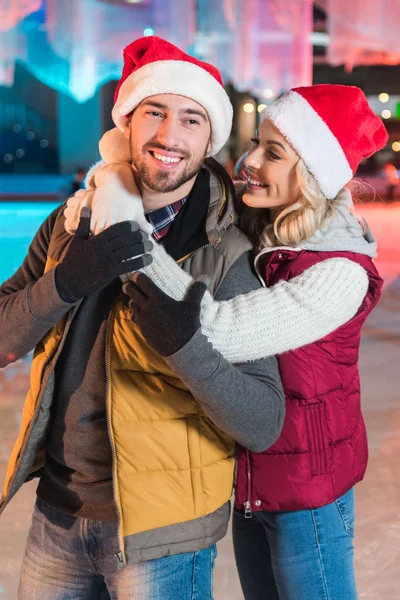 Bella felice giovane coppia in cappelli di Babbo Natale abbracciando sulla pista di pattinaggio — Foto stock