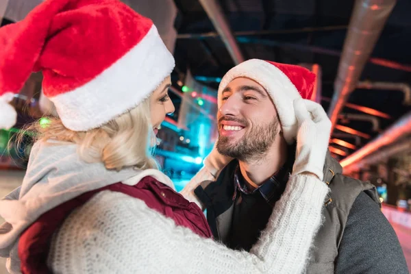 Feliz joven pareja en santa sombreros sonriendo el uno al otro en la pista - foto de stock