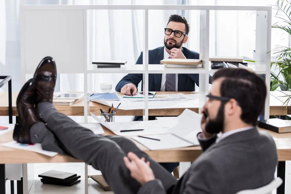 Pensativo hombre de negocios tratando de trabajar cuando colega hablando por teléfono inteligente con las piernas en la mesa - foto de stock