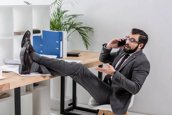 Sorprendido hombre de negocios hablando por teléfono inteligente con las piernas en la mesa - foto de stock