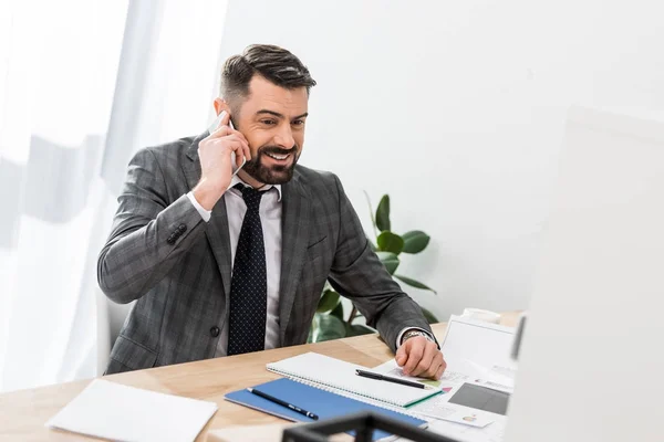 Homme d'affaires souriant parlant par smartphone à la table dans le bureau — Photo de stock