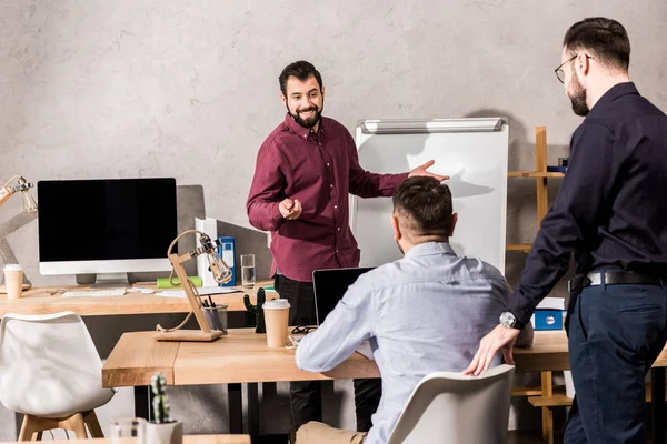 Uomo d'affari sorridente che mostra qualcosa ai colleghi a flipchart in ufficio — Foto stock