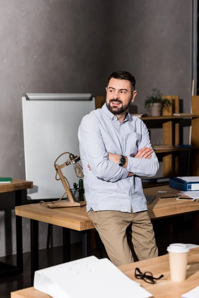 Homme d'affaires souriant avec les mains croisées appuyé sur la table dans le bureau et détournant les yeux — Photo de stock