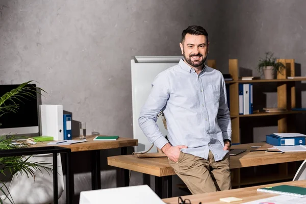 Lächelnder Geschäftsmann lehnt sich im Büro auf den Tisch und blickt in die Kamera — Stockfoto