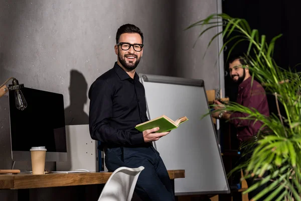 Lächelnder Geschäftsmann steht mit Buch im Büro und blickt in die Kamera — Stockfoto