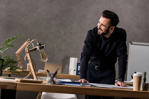 Arquitecto sonriente apoyado en la mesa de trabajo y mirando hacia otro lado - foto de stock