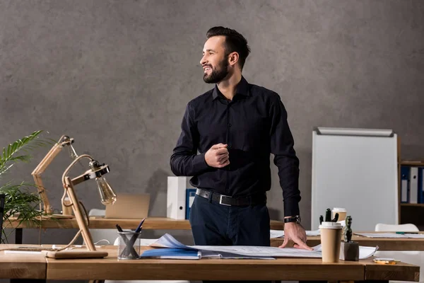 Smiling architect standing near working table and looking away — Stock Photo