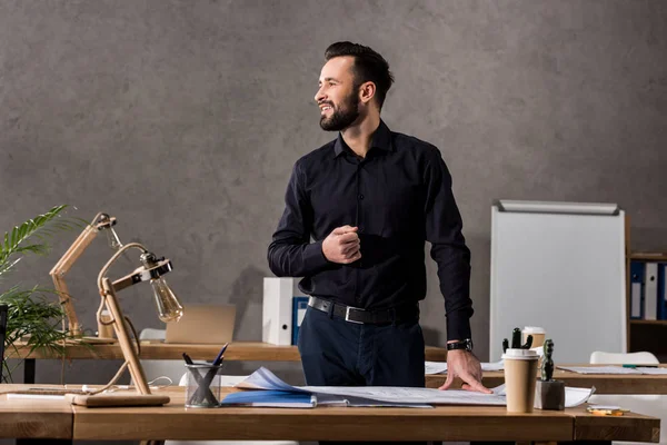 Arquiteto sorrindo de pé perto da mesa de trabalho e olhando para longe — Fotografia de Stock