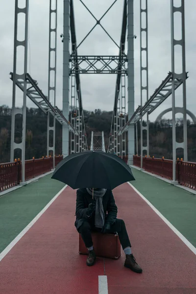 Stylish man with umbrella sitting on suitcase on pedestrian bridge — Stock Photo