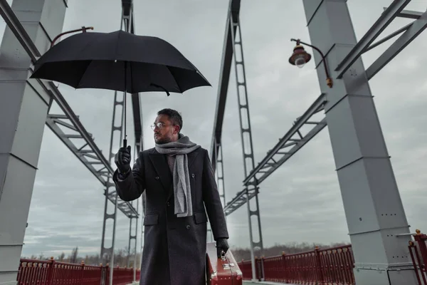 Lonely adult man with umbrella and luggage walking by bridge — Stock Photo