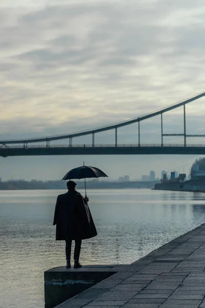Homme élégant avec parapluie debout sur la rive de la rivière — Photo de stock